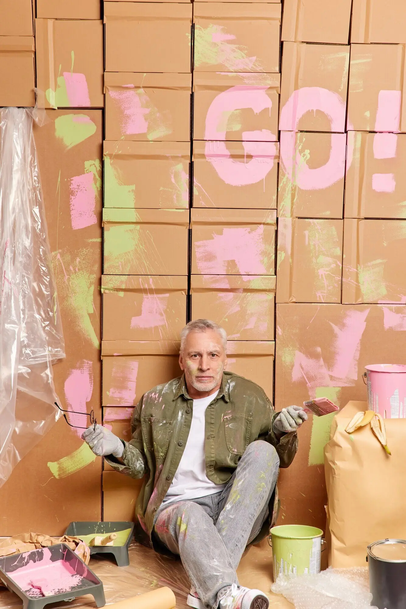 Innenaufnahme eines grauhaarigen Mannes, der zögernd mit den Schultern zuckt, umgeben von einem Farbeimer und Reparaturwerkzeugen; er sitzt auf dem Boden, nimmt die Brille ab und trägt legere Kleidung. Bau, Umbau oder Renovierung.